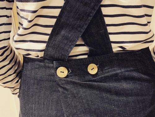 An image of the back of a denim dress where the straps cross over and are closed with wooden badges
