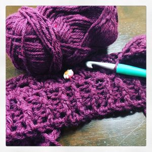 A purple ball of yarn and some crochet with a crochet hook that will become a gin o'clock cardigan 