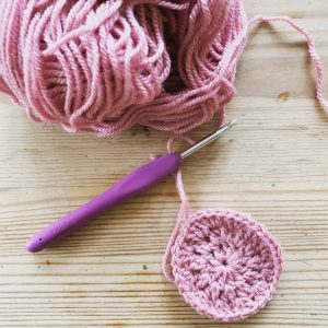 A purple crochet hook with a pink ball of yarn and the beginnings of a granny square